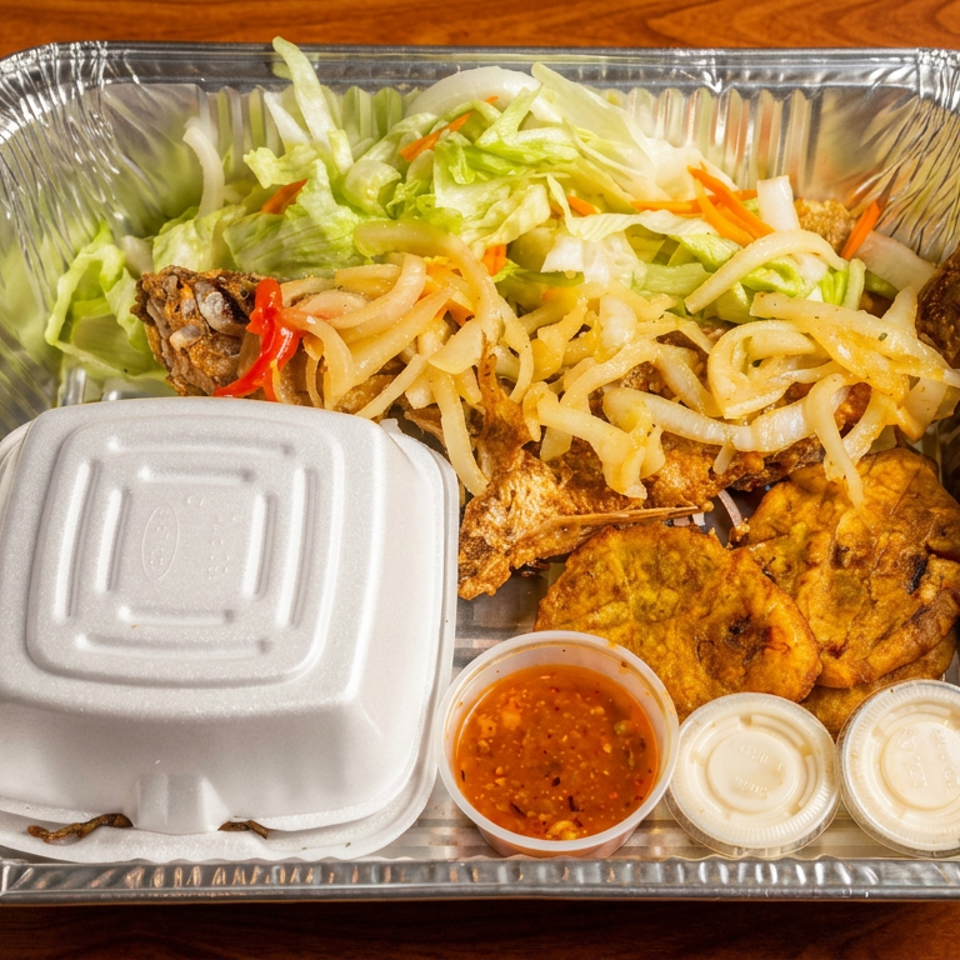Aluminum takeout tray filled with fried fish, shrimp fritters, noodles, salad, rice, and dipping sauces on a wooden table.